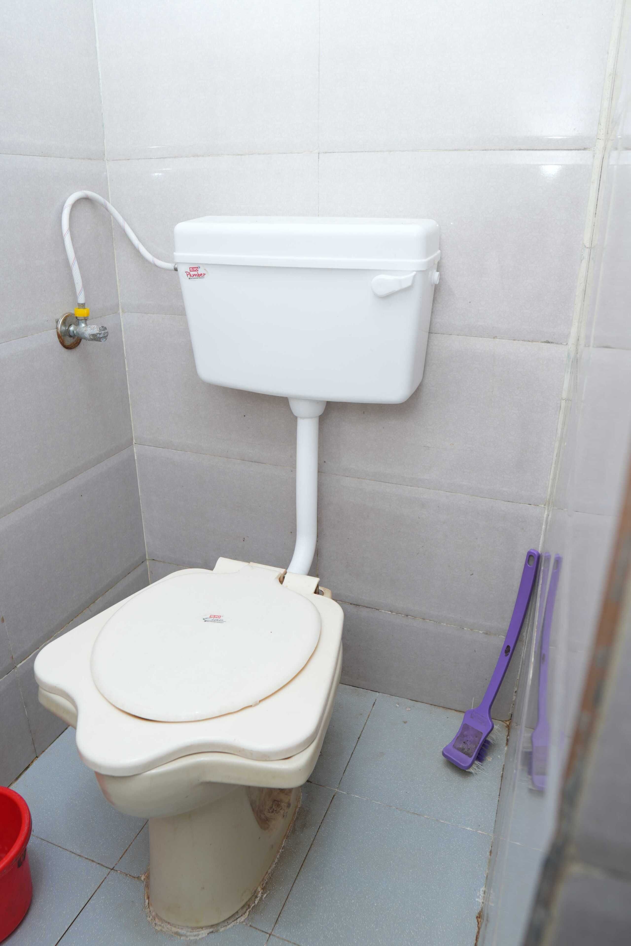 Inodoro blanco con tanque y asiento, un cubo rojo y una herramienta de limpieza morada en un baño con azulejos.