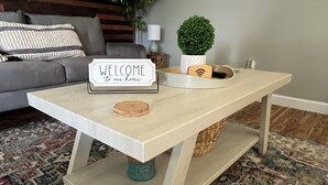 Een moderne woonkamer met een lichtkleurige salontafel, een welkomstbord, een kleine potplant en een decoratieve houten dienblad.