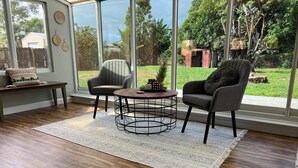 Een ruime kamer met een groot raam, een ronde salontafel, twee fauteuils, een bankje en een kamerkleed met dessin.