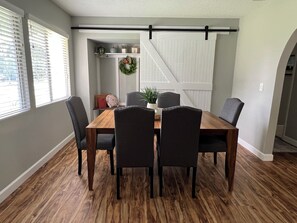 Een eetruimte met een houten tafel en zes stoelen, een decoratieve krans aan de muur en een schuifdeur in boerderijstijl.
