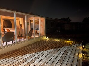 Een houten terras met buitenverlichting, een glazen ruimte met een eethoek en uitzicht op het omliggende landschap.