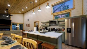 A modern kitchen with a central island, stainless steel appliances, and a dining area with wooden chairs and a table set for four. - Sage Creek: Secluded Family Getaway | Large Deck (Broken Bow)