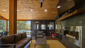 A covered outdoor seating area with a fireplace, wooden ceiling, and large windows. - Sage Creek: Secluded Family Getaway | Large Deck (Broken Bow)