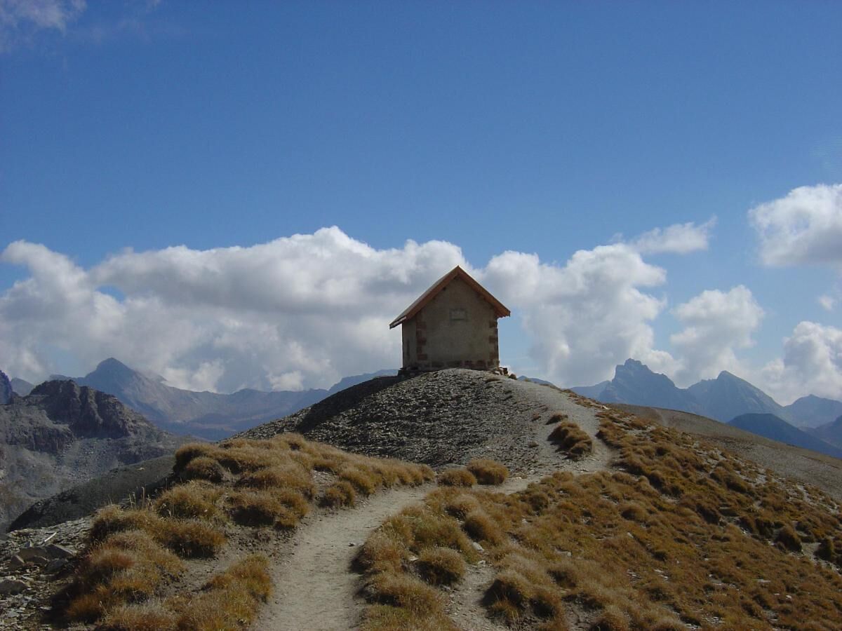 A small stone building with a red roof on a hilltop, surrounded by mountains and a clear sky.