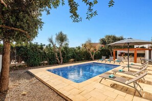 A rectangular swimming pool with a tiled edge, surrounded by a pebble-lined deck. There are lounge chairs and a pergola with an umbrella.