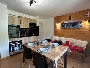 A dining area with a wooden table set for four, a kitchen with black appliances, and a wooden wall with a mountain painting.