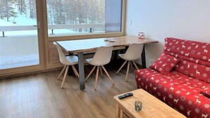 A room with a wooden dining table and chairs, a red sofa with a festive pattern, and a large window offering a view of a snowy landscape. - 20m from ski slopes, 3rd floor, balcony, parking, 50m², Vars (Vars)