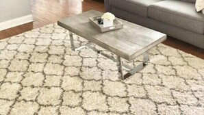 Una sala de estar con un sofá gris, una mesa de centro de madera y un tapete hexagonal. Hay un ventilador de techo, una zona de comedor con sillas blancas y una cocina al fondo.