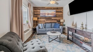 A living room with a grey sofa, a coffee table, a ceiling fan, and a wooden wall panel. - Pool View King Suite #108 (New Smyrna Beach)