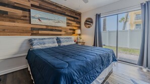 A bedroom with a large bed, wooden headboard, a ceiling fan, a beach painting, and a view of the outside. - Pool View King Suite #108 (New Smyrna Beach)