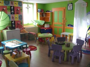 Een vrolijk ingerichte kinderspeelruimte met diverse tafels en stoelen, een boekenkast en een radiator.
