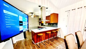 A modern kitchen with a central island, bar stools, wooden flooring, and a large screen displaying a streaming service interface. - King beds Hot Tub Arcade Luxury Showers (Kernersville)