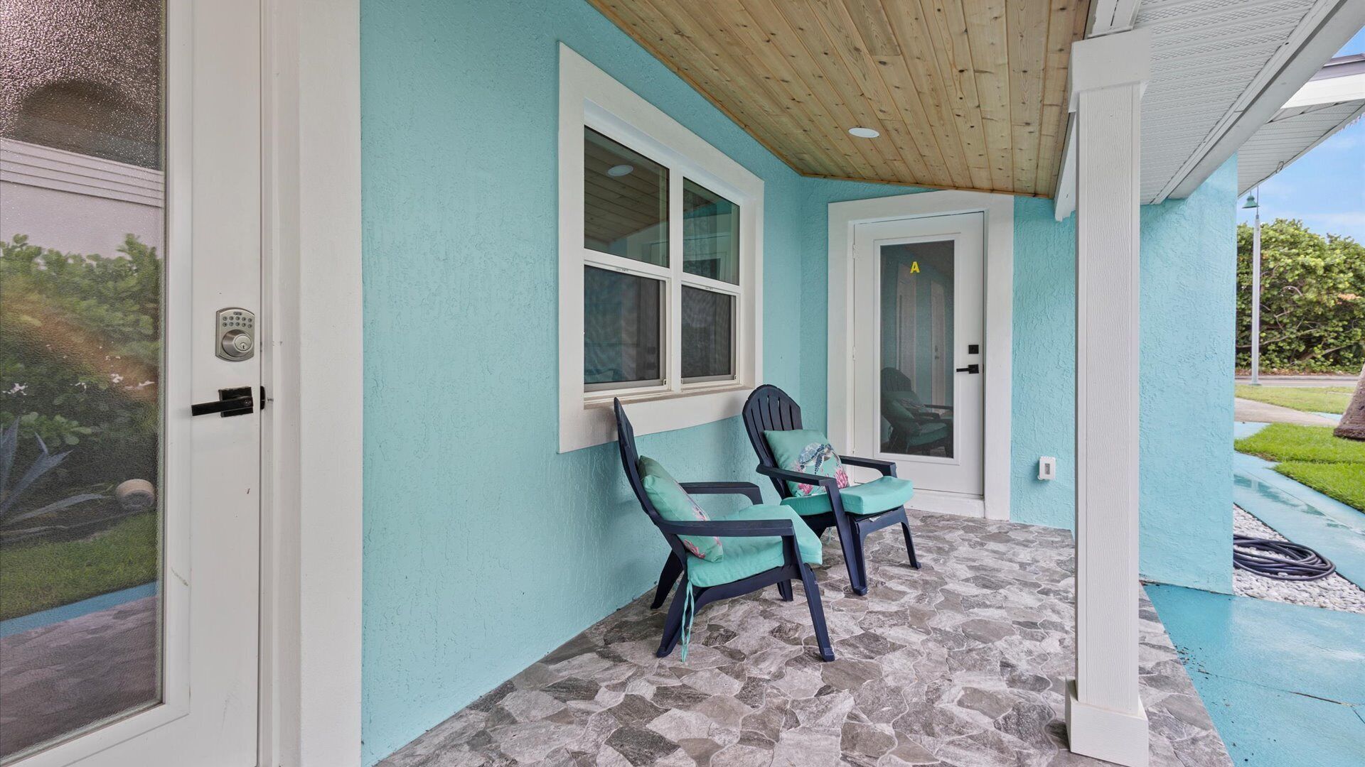A covered porch with two chairs and a table, a door with a keypad lock, and a window with a view of greenery.