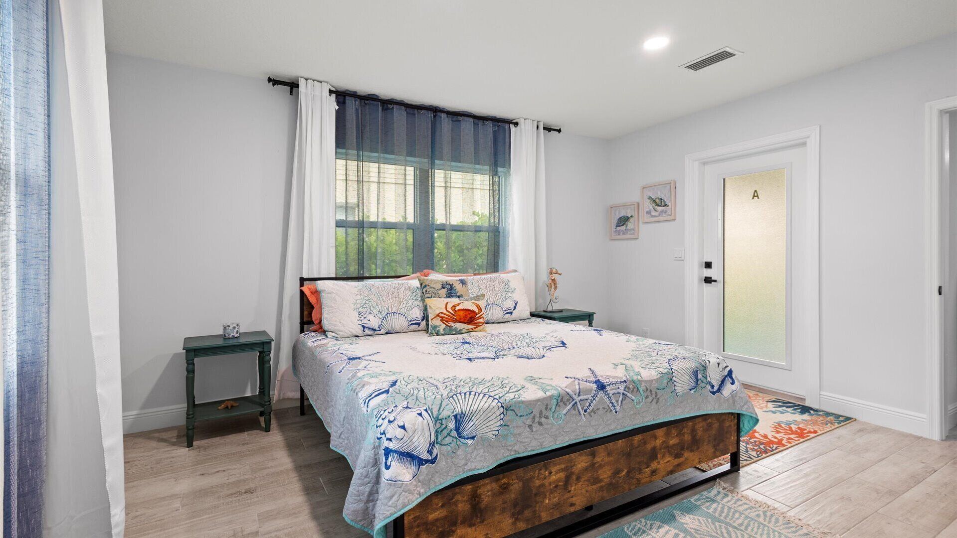 Un dormitorio con cama, mesitas de noche, una ventana con cortinas, una puerta y un pequeño adorno en la pared.