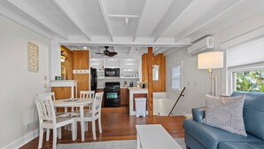Una cocina moderna con armarios blancos, un refrigerador negro y un microondas. Hay una zona de comedor con sillas blancas y una mesa, y un sofá azul con un cojín estampado.
