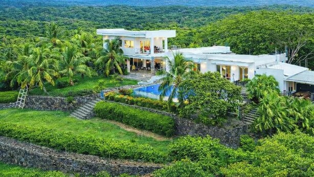 Une maison moderne à deux étages avec une piscine, entourée d’une végétation luxuriante et avec une montagne en toile de fond.