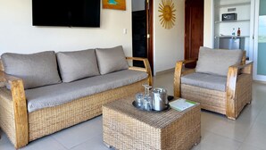 Un salon comprenant un canapé, des fauteuils, une table basse avec des verres et une bouilloire, un téléviseur, un tableau et un élément de décoration représentant un soleil.