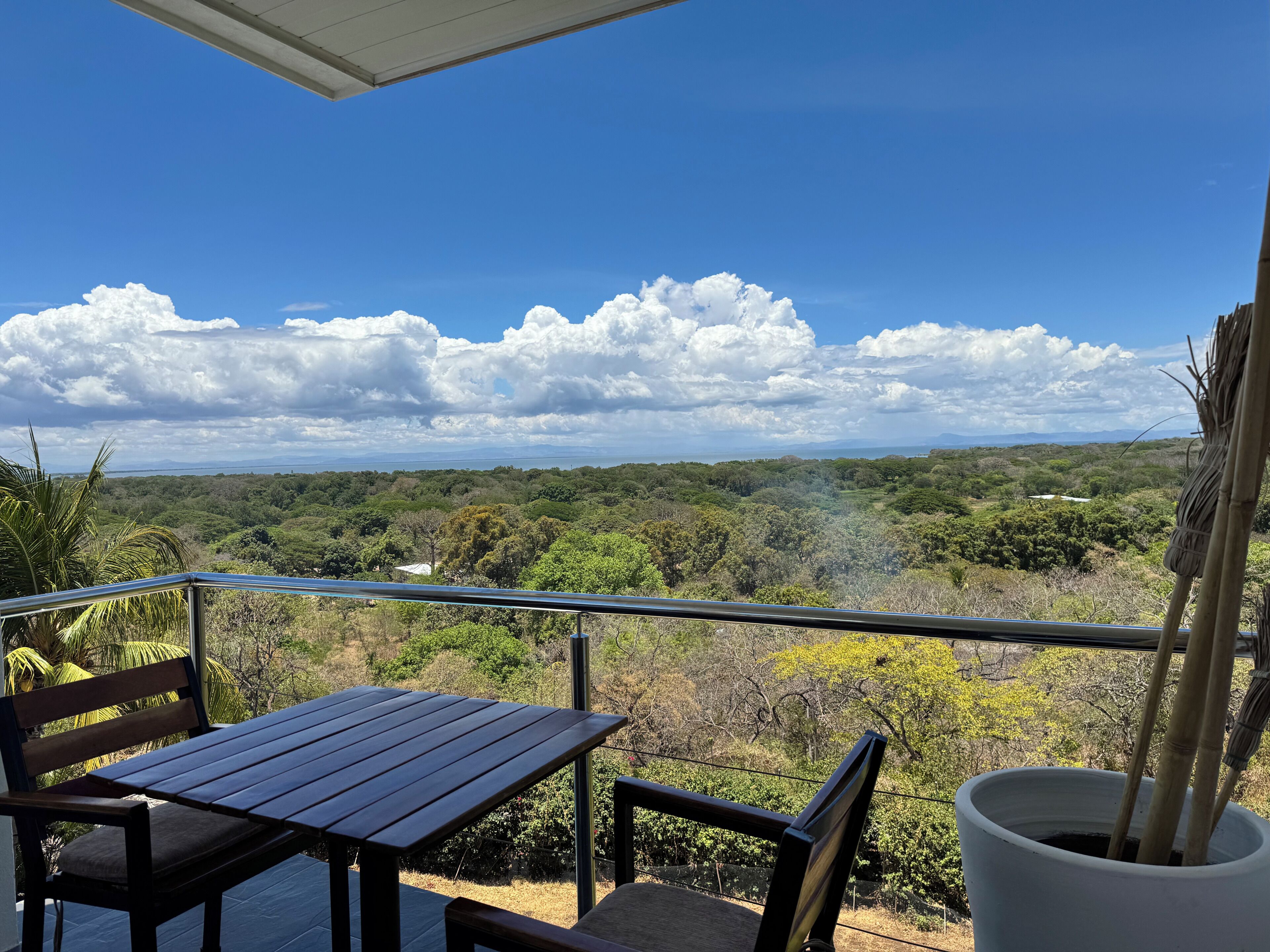 A balcony with a table and chairs, overlooking a lush landscape with trees and a distant mountain range.
