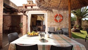 An outdoor patio with a table set for dining, surrounded by wicker chairs. A thatched roof provides shade, and a lifebuoy hangs on the wall. - H2933 Finca Tia in Pollenca (Pollenca)