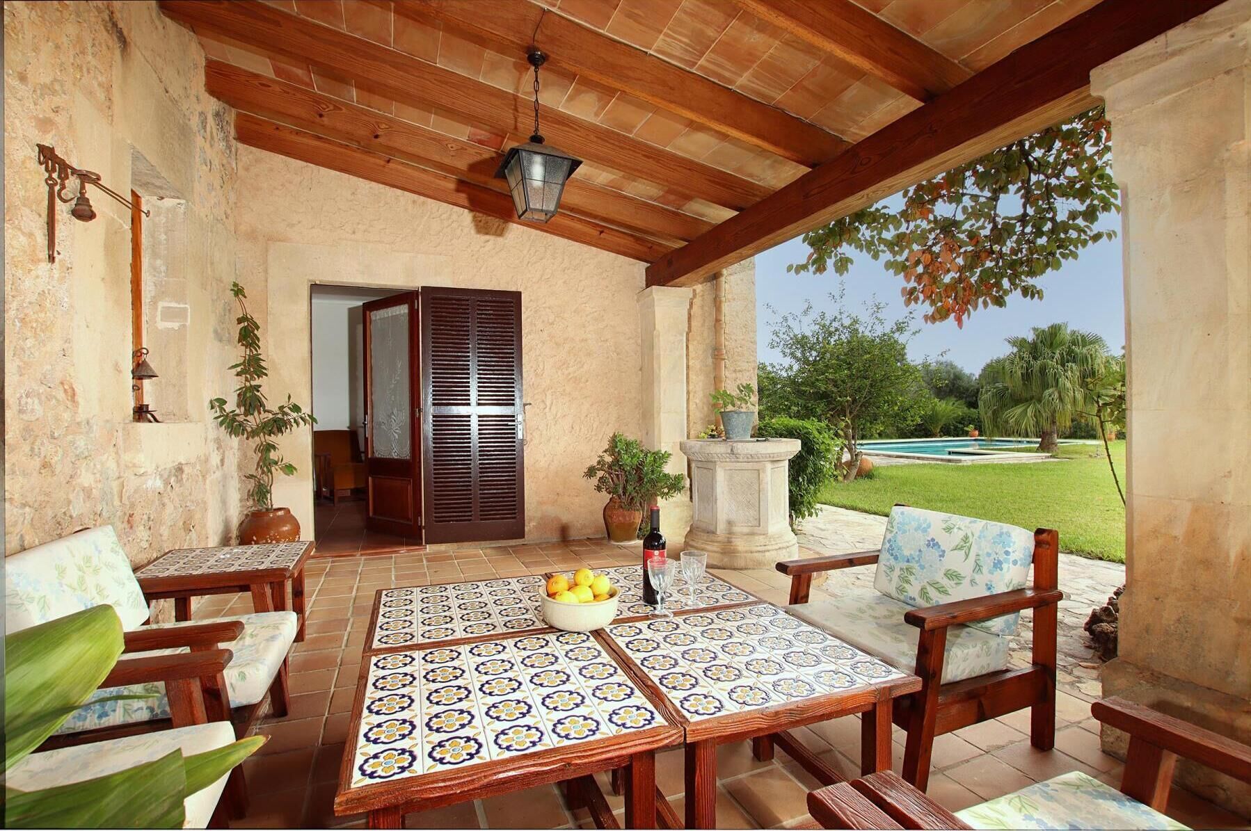 A covered patio with a tiled table, wooden chairs, a potted plant, and a view of a swimming pool and greenery.