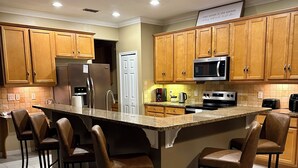 Eine moderne Küche mit Holzausstattung, einem Edelstahlkühlschrank, einer Mikrowelle und einem eingebauten Backofen. Vier Barhocker stehen an der Kücheninsel.