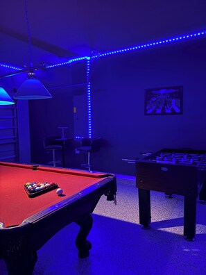 A pool table with balls, a foosball table, and a framed picture on the wall.