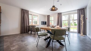 Una habitación amplia con una gran mesa de madera, seis sillas, un televisor y ventanas grandes con cortinas.