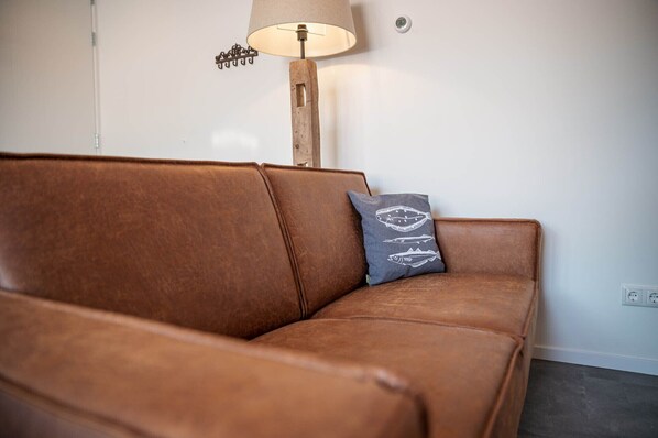 A brown leather sofa with a gray pillow featuring fish designs, a wooden floor lamp, and a white wall with a power outlet. - De Witte Brugh- 't Baken (Buren)