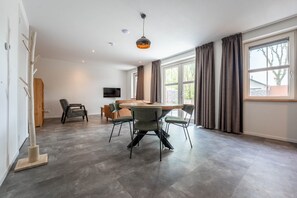 A spacious room with a wooden dining table, four chairs, a flat-screen TV mounted on the wall, and large windows with curtains. - De Witte Brugh- De Miëde (Buren)