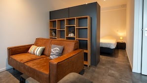 A modern room with a brown leather sofa, a built-in wooden shelving unit, a bed with white bedding, and a nightstand with a lamp. - De Witte Brugh- 't Fennoat (Buren)