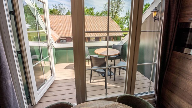 A covered outdoor deck with a table and chairs, a sliding glass door, and a view of a house with a red roof. - De Witte Brugh - Nijlânsrèèd (Buren)