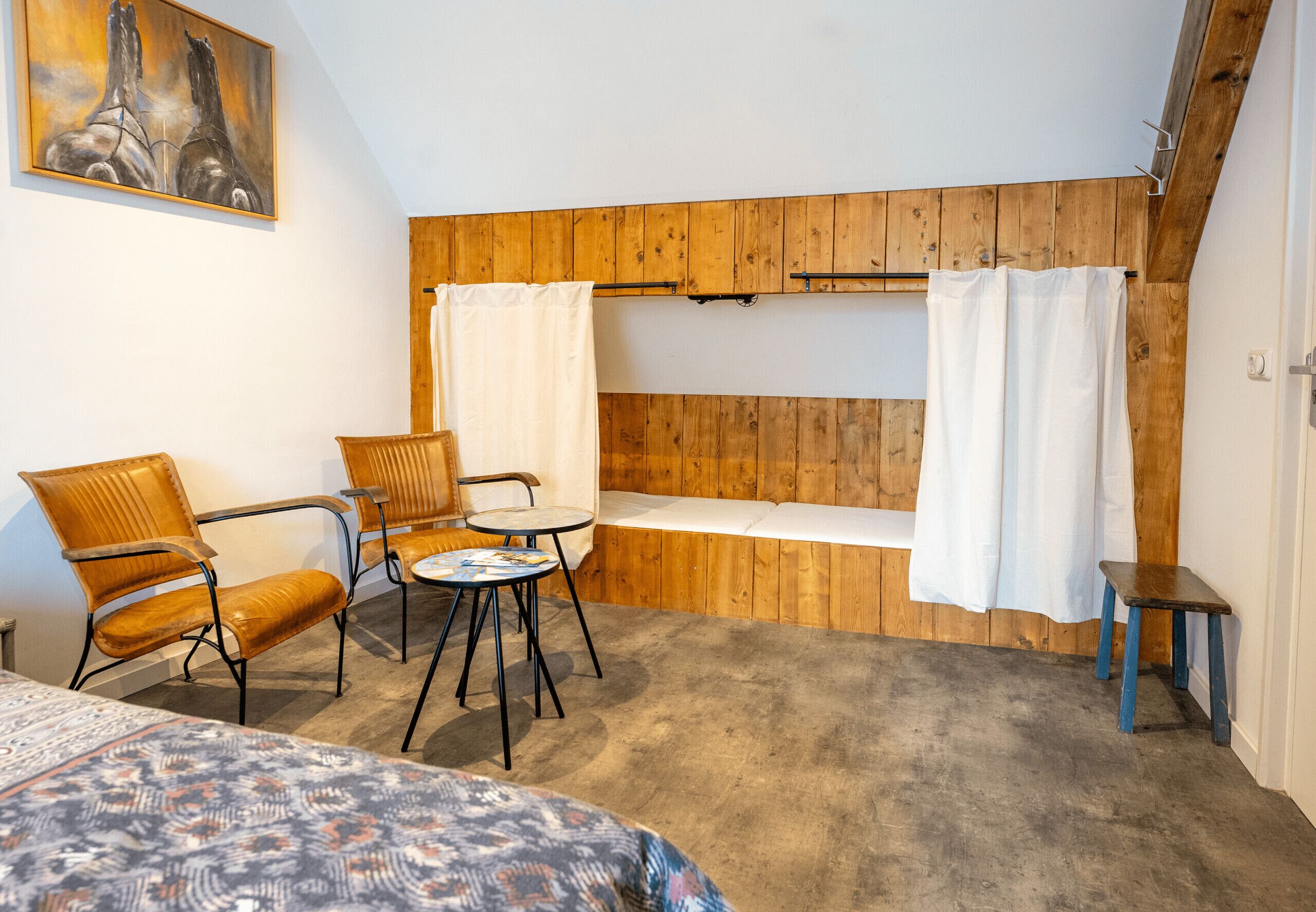 Ett rum med ett sluttande tak av trä, en säng med ett mönstrat överkast, två trästolar, ett litet runt bord och en bänk med en vit gardin.