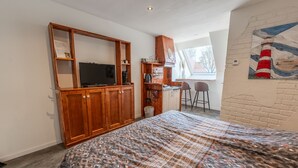 Ett sovrum med en säng, en garderob i trä, en tv, ett litet kök och en tavla på väggen.