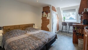 Een slaapkamer met een groot bed, een houten hoofdbord, een bakstenen muur, een raam en een kitchenette met krukjes.