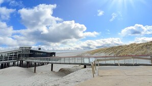 A beachside building with a glass facade and a name sign, a wooden boardwalk, and sandy dunes in the background. - De Witte Brugh - Studio 4 (Buren)