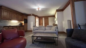 A living room with a sofa, a red armchair, a coffee table, and a kitchen area with wooden cabinets and a refrigerator.