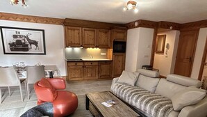 A living room with a sofa, a coffee table, and a red chair. There is a kitchen area with wooden cabinets and a microwave.