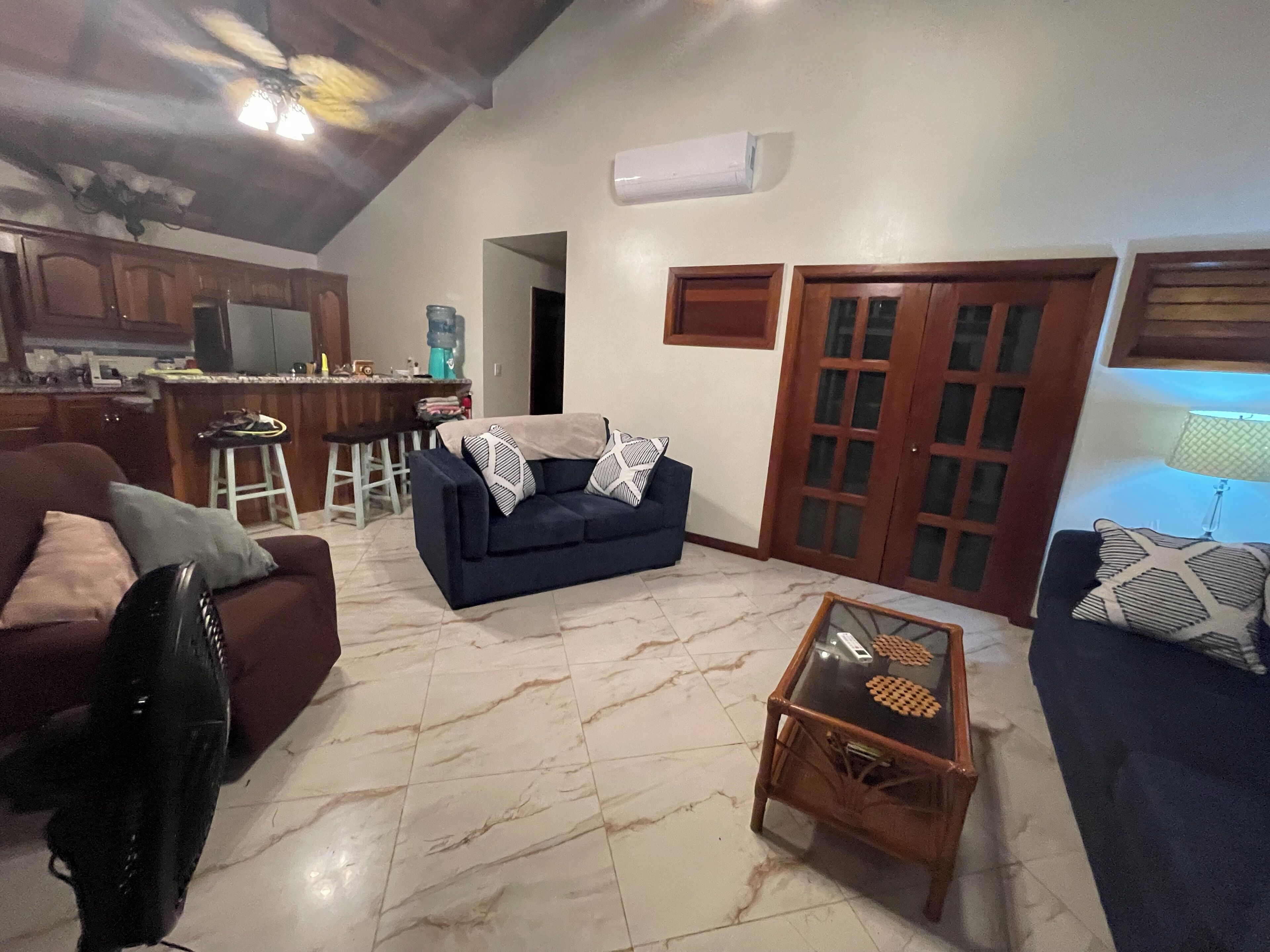 A living room with a sofa, a coffee table, and a TV. There is a kitchen area with a water dispenser and a bar stool. The room has a tiled floor and wooden doors.