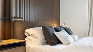 Una cama bien hecha con ropa de cama blanca y gris, un cabecero de madera y una mesita de noche con una lámpara.