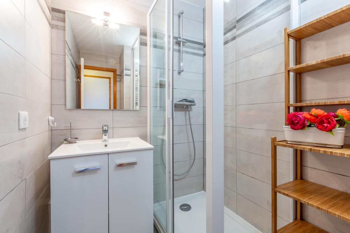 A modern bathroom with a glass shower enclosure, a white vanity with a mirror, and a wooden shelf with flowers.
