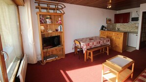 Una habitación compacta con un mueble bajo de madera para el televisor, una mesita, un banco y una zona de kitchenette.