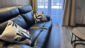 Ein modernes Wohnzimmer mit einer schwarzen Ledersofa, zwei gemusterten Kissen, einem Holztisch und Blick ins Freie durch große Fenster.