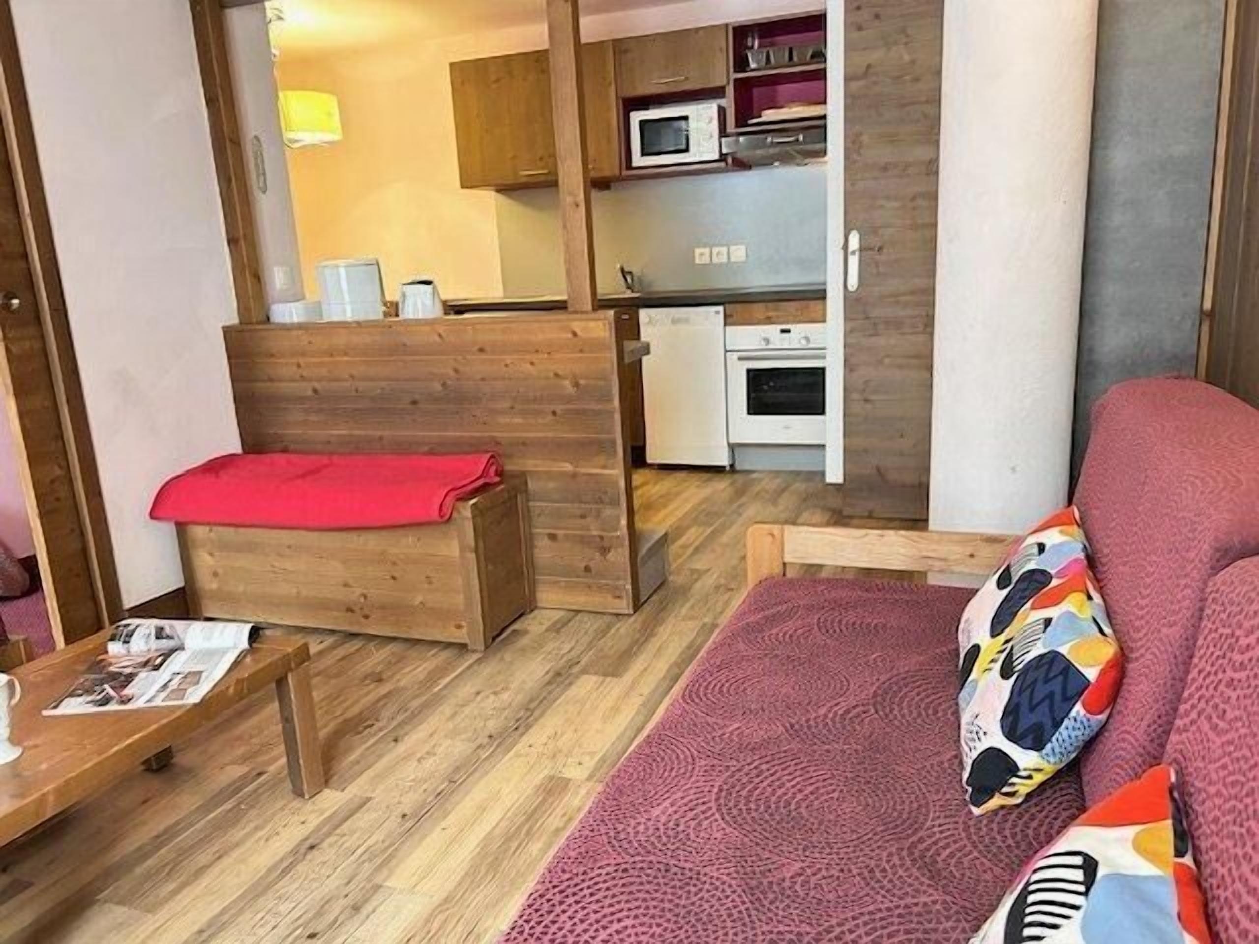 Ein kleines Zimmer mit einer hölzernen Sitzbank, einem Sofa mit bunten Kissen, einem kleinen Tisch mit einer Zeitung und einer Küchenzeile im Hintergrund.