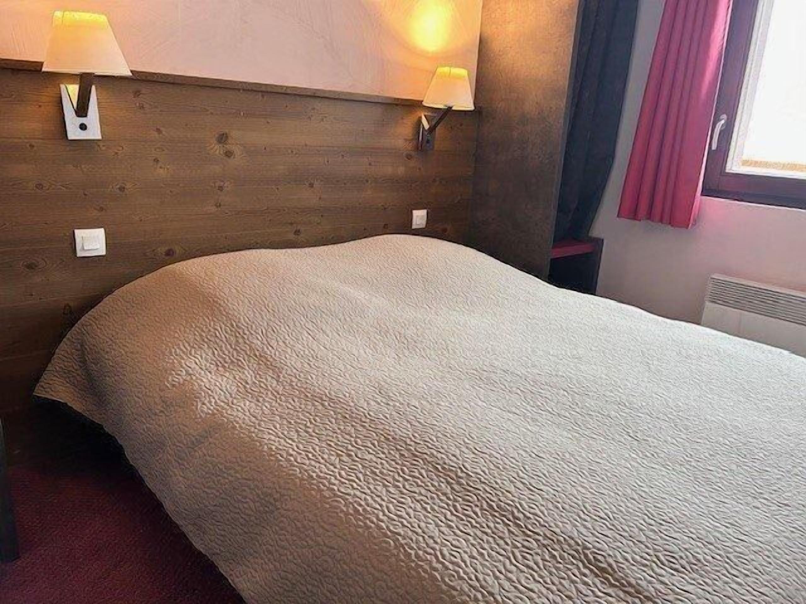 Hotellihuone, jossa on puinen sängynpääty, kaksi seinään kiinnitettyä lamppua, kuvioitu viltti sängyllä ja ikkuna, jossa on verhot.
