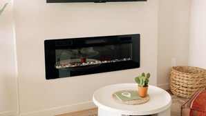 Een woonkamer met een flatscreen televisie boven een open haard, een ronde witte salontafel met een cactus, en een geweven mand.