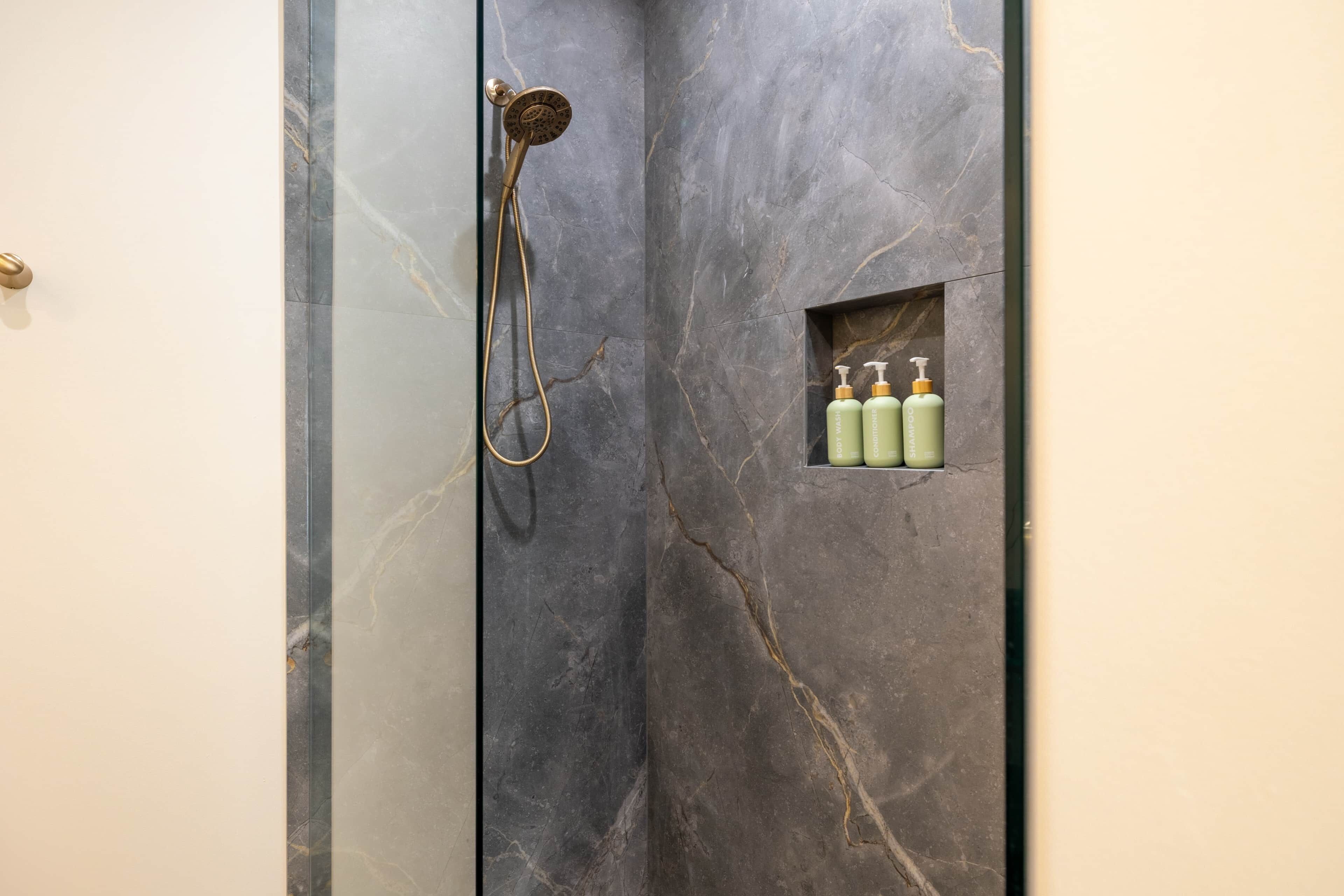 Ein Badezimmer mit Marmorwand, einer Dusche inklusive Handbrause und einem Ablagebereich mit drei Flaschen.