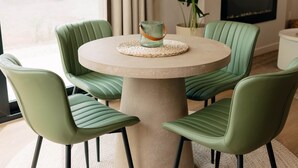 Een ronde tafel met vier groene stoelen, een geweven placemat, een potplant en een flatscreen televisie aan de muur.