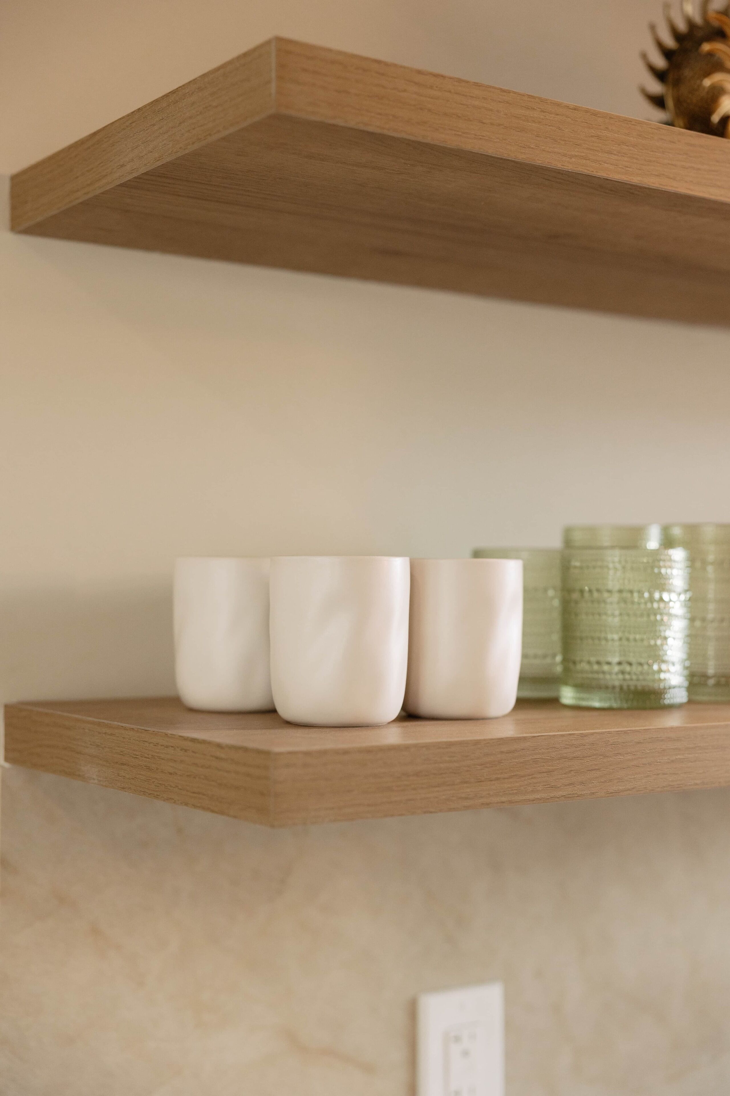 A wooden shelf with three white ceramic cups and a green glass container.