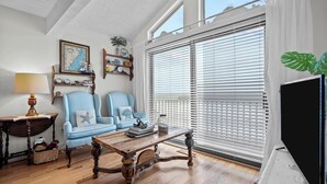 Un salon chaleureux avec une table basse en bois, deux fauteuils à motifs, une table d’appoint avec une lampe, une bibliothèque et une grande fenêtre avec des stores.