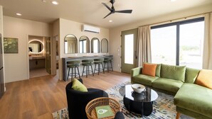 A modern living room with a green sofa, black armchair, and a coffee table. There is a kitchen area with a bar and stools, and a large window with curtains. - Vega 02: A spacious, 3-bedroom desert oasis stay, Vega #2 (Pinal County)